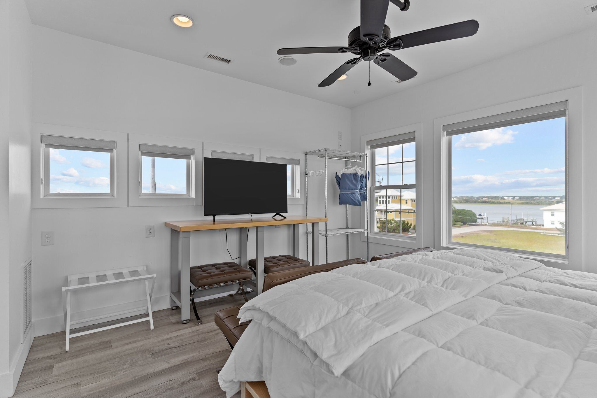 A bedroom with a large bed, a TV on a stand, a ceiling fan, and a view of a body of water.