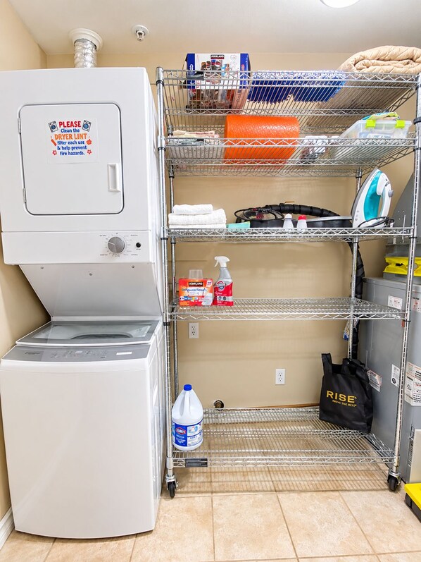 A laundry area with a washing machine, a metal shelf with folded towels, cleaning supplies, and a water bottle.