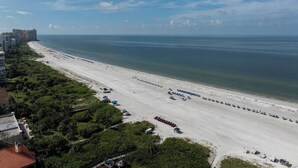 A beach with rows of sun loungers and umbrellas, a clear sky, and buildings in the background. - Sea Suite (Marco Island)
