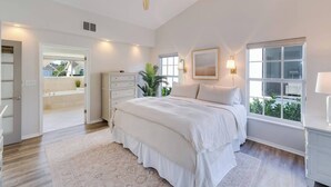 A bedroom with a large bed, a ceiling fan, a window with a view, and a nightstand with a lamp. - Sea Suite (Marco Island)