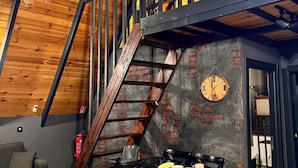 Una habitación estilo loft con techo de madera, paredes de ladrillo visto, una escalera de metal negro y un comedor con mesa y sillas.
