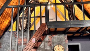 Habitación estilo loft con vigas de madera a la vista, pared de ladrillo y escalera de madera. Hay un reloj en la pared y una mesa con sillas.