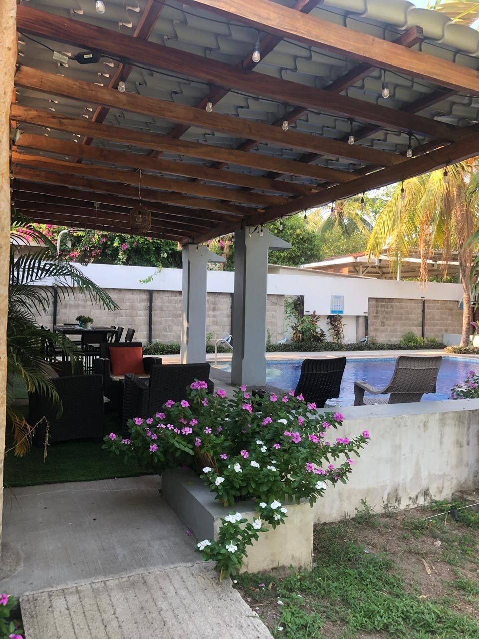 A covered outdoor seating area with a pool, surrounded by greenery and a concrete wall.