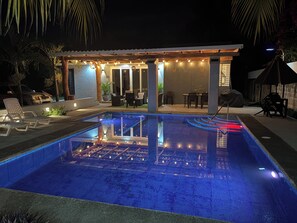 A swimming pool with a tiled deck and lounge chairs, surrounded by a covered patio area with tables and chairs.