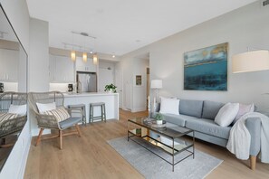 Una sala moderna con un sofá, una mesa de centro de vidrio, sillas de mimbre y una cocina con armarios blancos y un refrigerador.