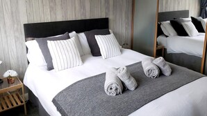 A neatly made bed with pillows and a blanket, a small wooden nightstand with a vase of flowers, and a mirror on the wall. - 3 bedroom accommodation in Conwy (Conwy)