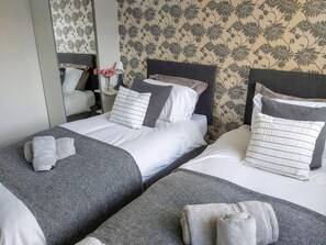 Une chambre d’hôtel avec deux lits, chacun doté de draps gris et d’oreillers blancs, une table de chevet avec une lampe et un papier peint à motifs floraux.