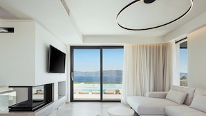 Ein modernes Wohnzimmer mit Flachbildfernseher, einer Couch, zwei runden Tischen und Blick auf Pool und Berge.