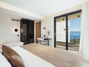 A modern bedroom with a bed, a chair, a small table, and a sliding glass door leading to a balcony with a view of the sea.