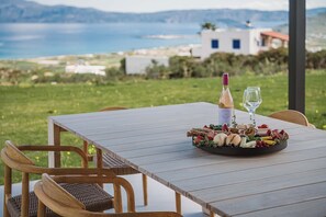 A wooden table with a fruit and cheese platter, a bottle of wine, and two glasses on a terrace with a scenic view of the sea and buildings.