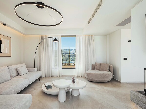 A modern living room with a sofa, round coffee table, and a chair, a large window with curtains, and a ceiling light fixture.