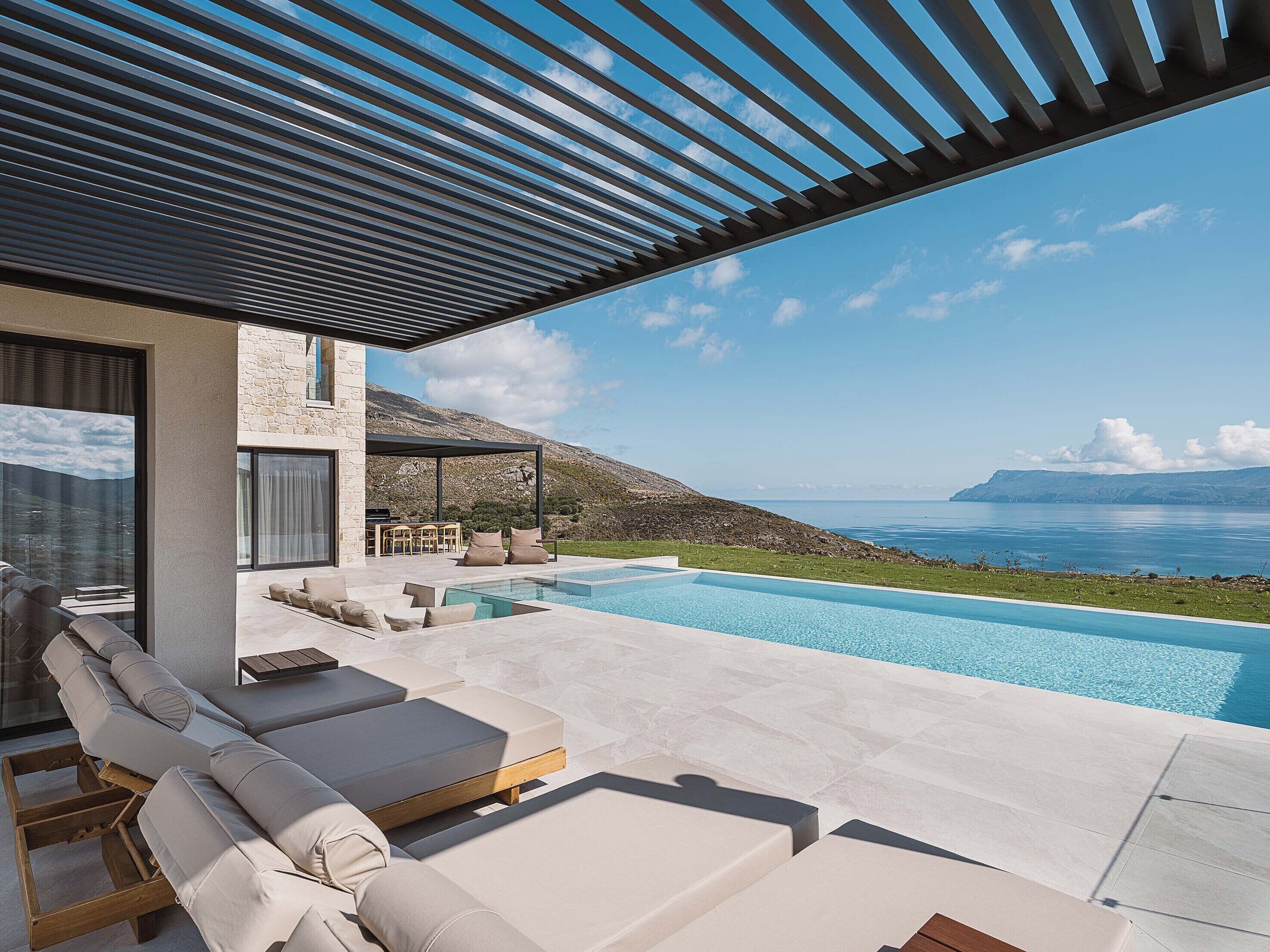 A modern outdoor pool area with lounge chairs and a stone building with a view of the sea.