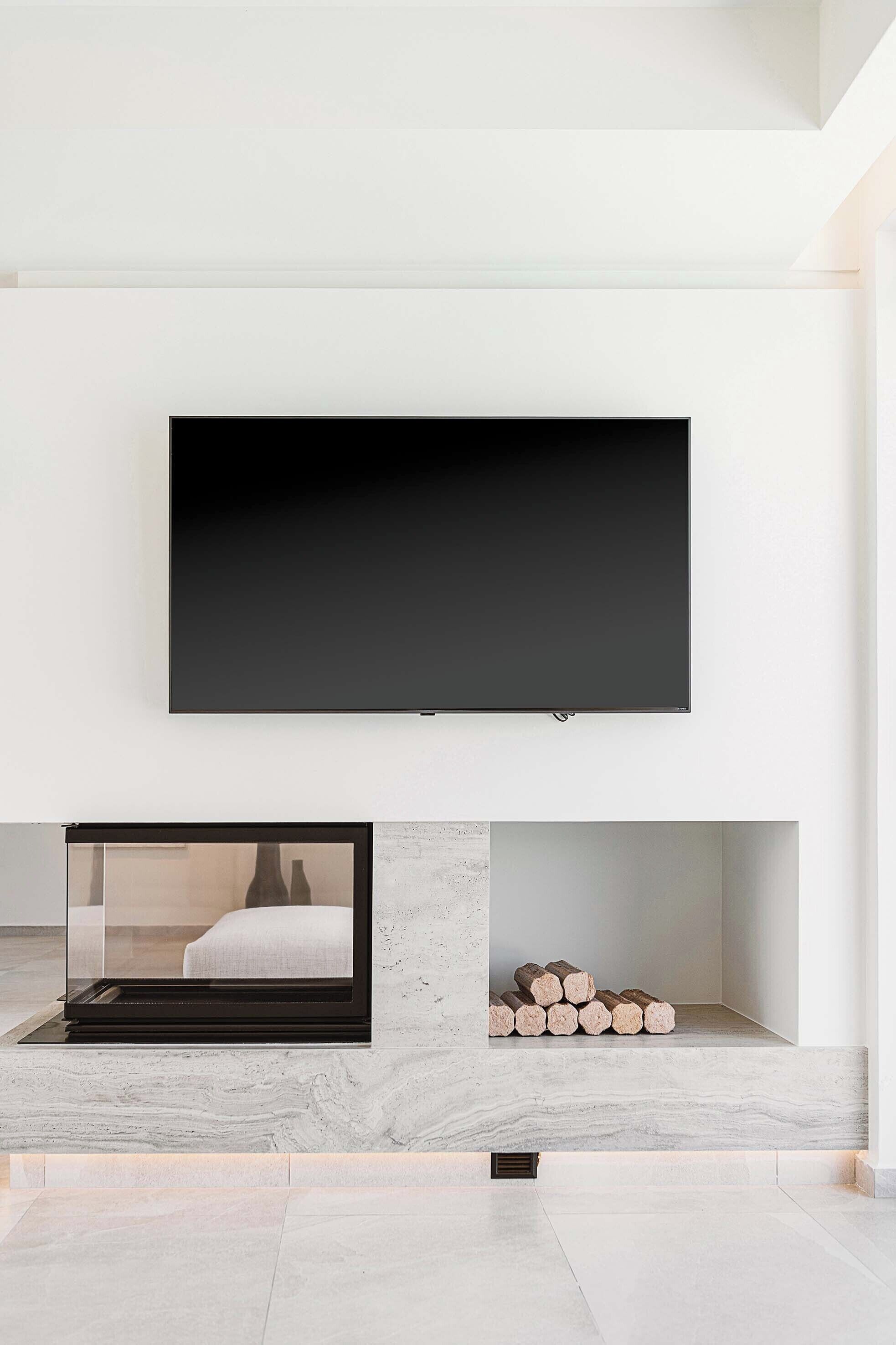 Un salon moderne avec un grand téléviseur à écran plat fixé au mur, une cheminée à foyer vitré à travers lequel on aperçoit un lit, et une pile de bois de chauffage sur l’étagère inférieure.