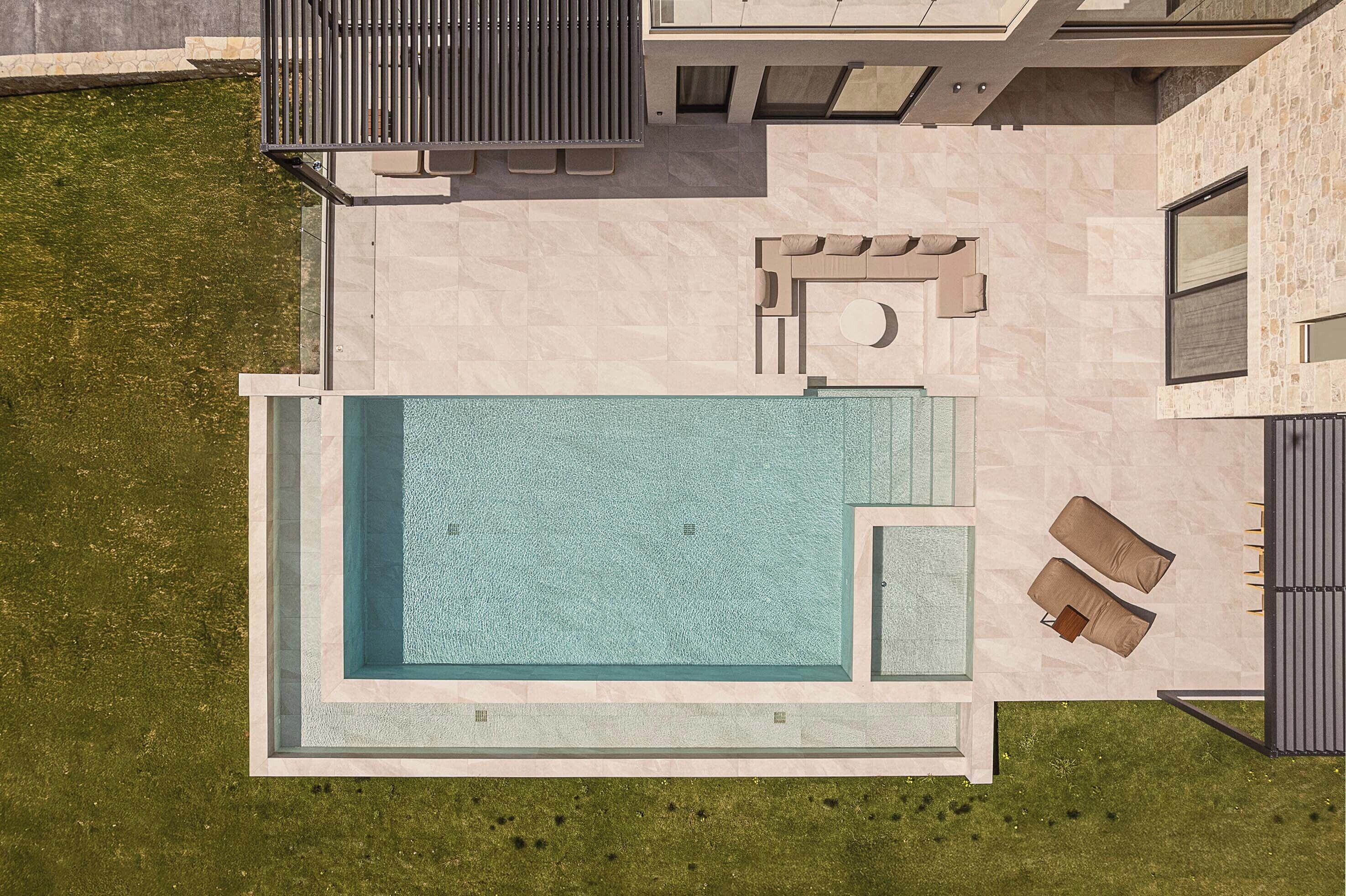 An aerial view of a modern house with a rectangular swimming pool, a patio with two lounge chairs, and a small outdoor seating area.