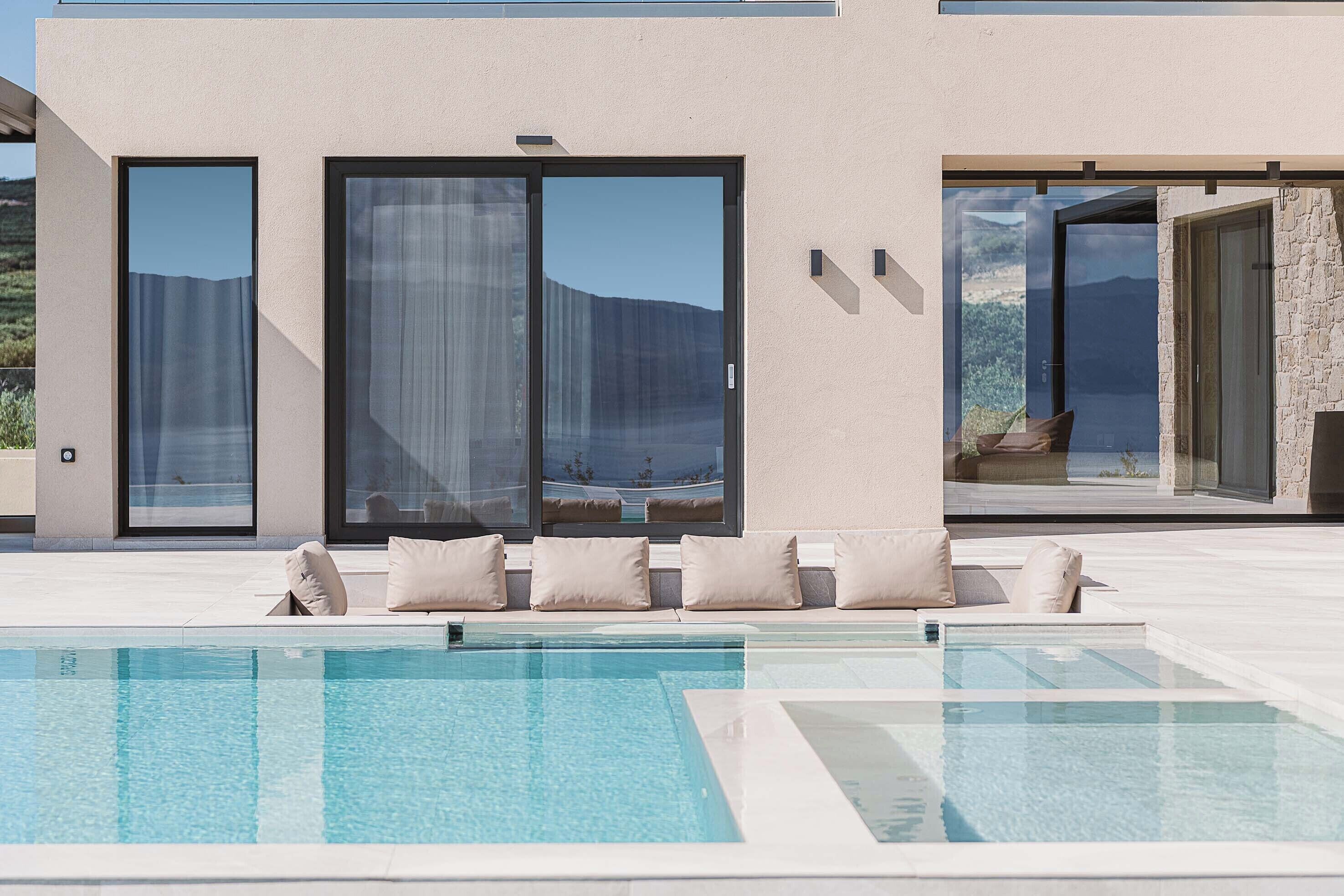 A modern pool area with a clear view of the surrounding landscape, a seating area with cushions, and large sliding glass doors.