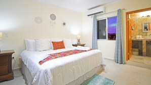 Un dormitorio con cama, mesita de noche, ventilador de techo, ventana con cortinas y baño visible a través de una puerta abierta.