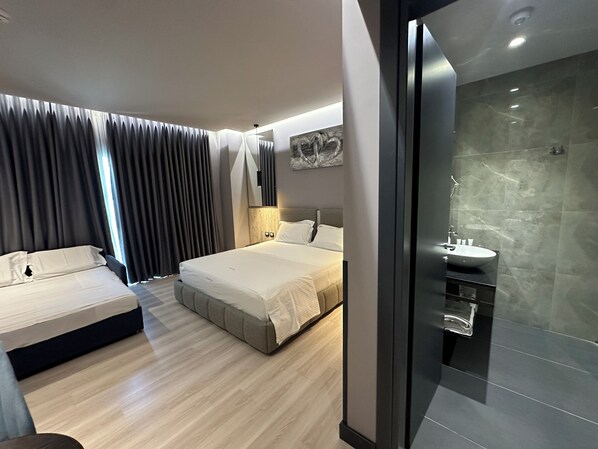 Una habitación de hotel moderna con una cama grande, un sofá independiente, un baño con ducha de vidrio y un amplio ventanal con cortinas.