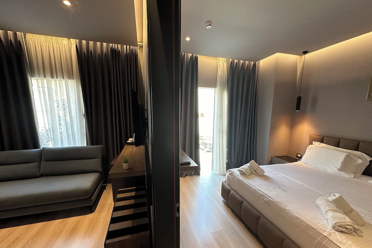 A modern hotel room with a sofa, a bed with neatly folded towels, a small table with a plant, and a view of the outdoors through large windows.