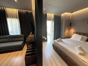 Una habitación de hotel moderna con sofá, cama con toallas ordenadas, una mesita con planta y vistas al exterior a través de amplios ventanales.