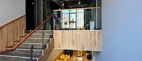 Un interior moderno con una escalera que baja a un nivel inferior, un comedor con sillas amarillas y vistas a edificios exteriores.