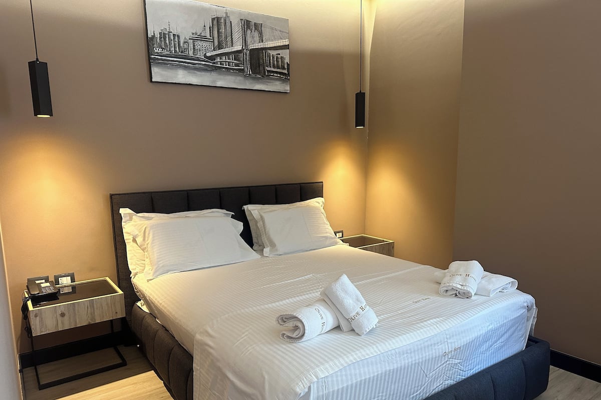 A hotel room with a large bed, two bedside tables, a nightstand, a wall-mounted painting, and a hanging light fixture.