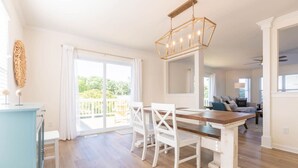 A dining area with a wooden table and white chairs, a chandelier, and a view of the outdoors through large sliding glass doors. - Tidal Watch 1-Master Suite w/ Ocean View (Kure Beach)