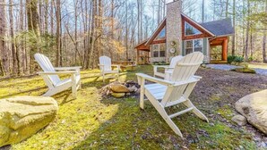 Eine Holzhütte mit einem Steinkamin, umgeben von einem Wald. Vor dem Haus befinden sich Adirondack-Stühle und eine Feuerstelle.
