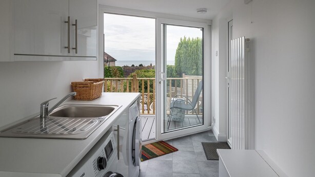 A modern kitchen with a built-in washing machine, a large window with a view of the sea, and a balcony with outdoor furniture. - Awel Fryn - Three Bedroom Cottage, Sleeps 6 (Hen Golwyn)