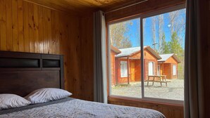 A wooden cabin with a bed, a window with a view of another cabin, and a wooden headboard. - Oceana Suites Rayen Lican-Ray (Villarrica)
