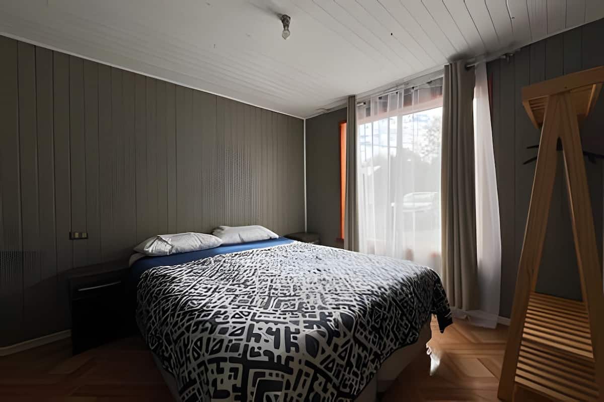 A bedroom with a bed, a wooden floor, a window with curtains, and a wooden wardrobe.
