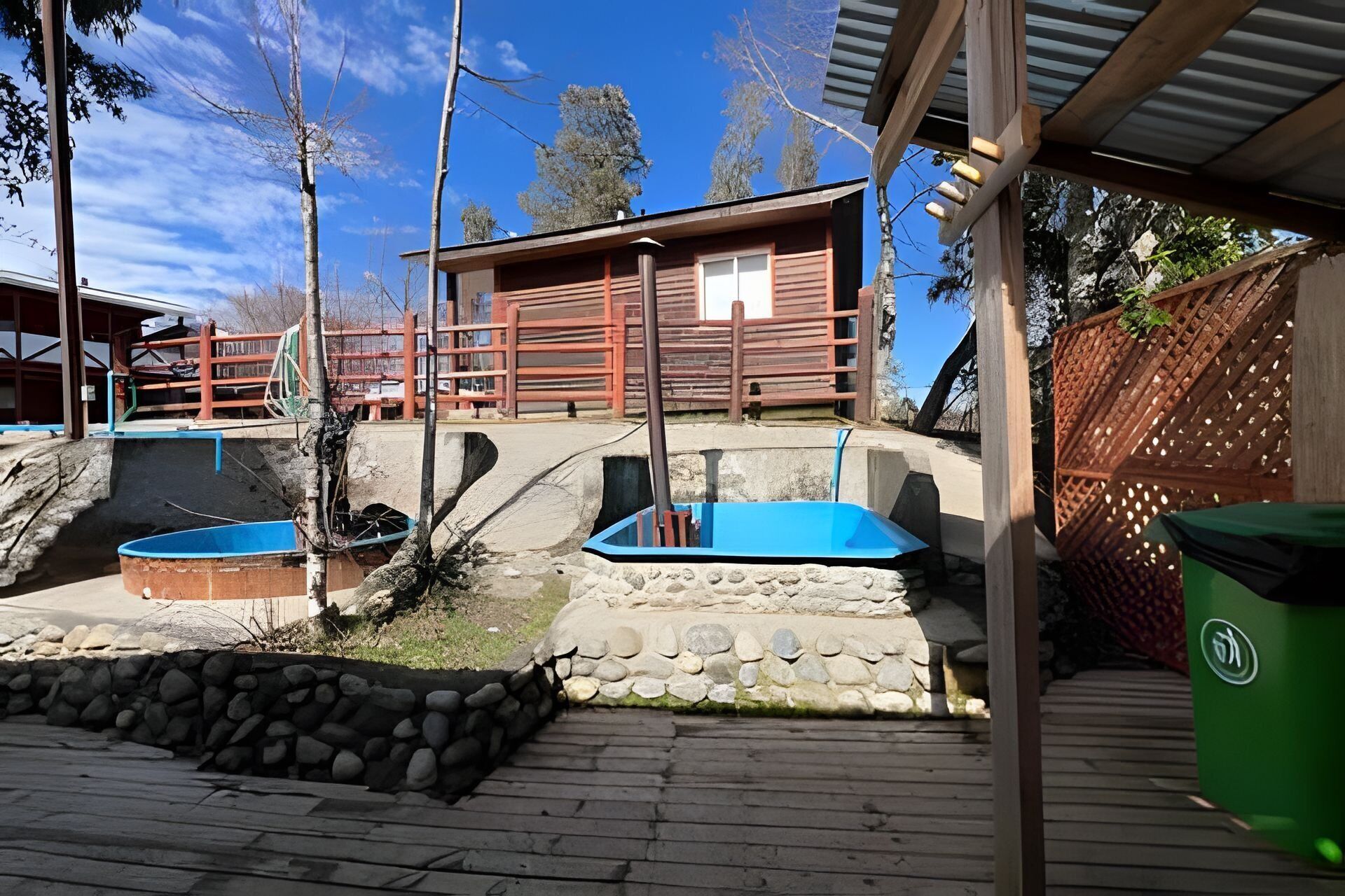 A wooden cabin with a stone pathway leading to a blue pool, surrounded by trees and a fenced area.