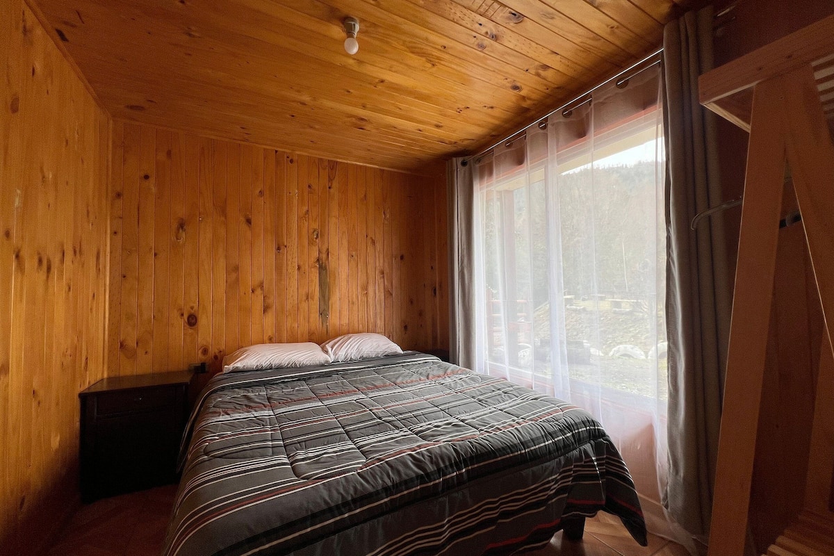 A wooden-paneled room with a bed, a window with curtains, and a wooden ceiling.