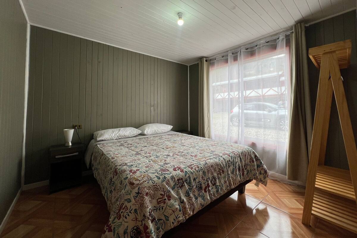 A bedroom with a bed, a nightstand, a wooden chair, a window with curtains, and a ceiling light.