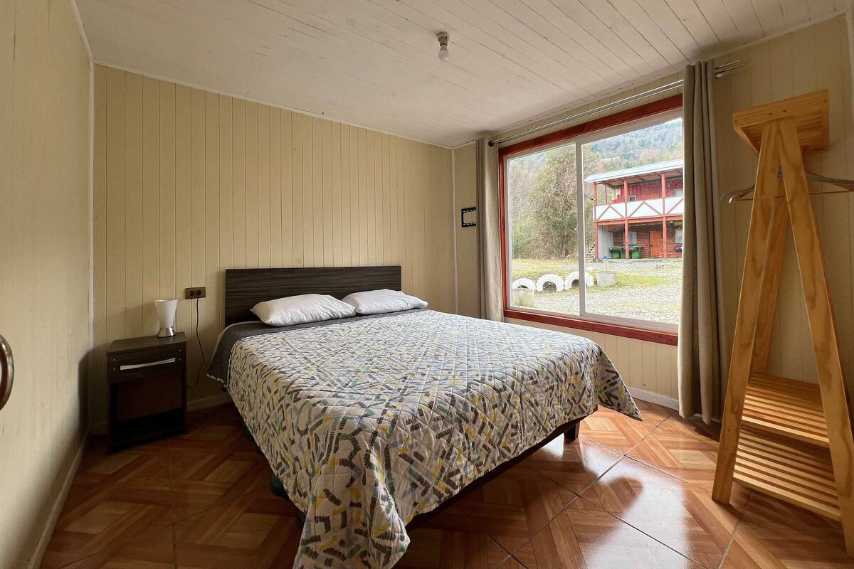 A bedroom with a bed, a nightstand, a lamp, a window with a view of a building, and a wooden ladder.