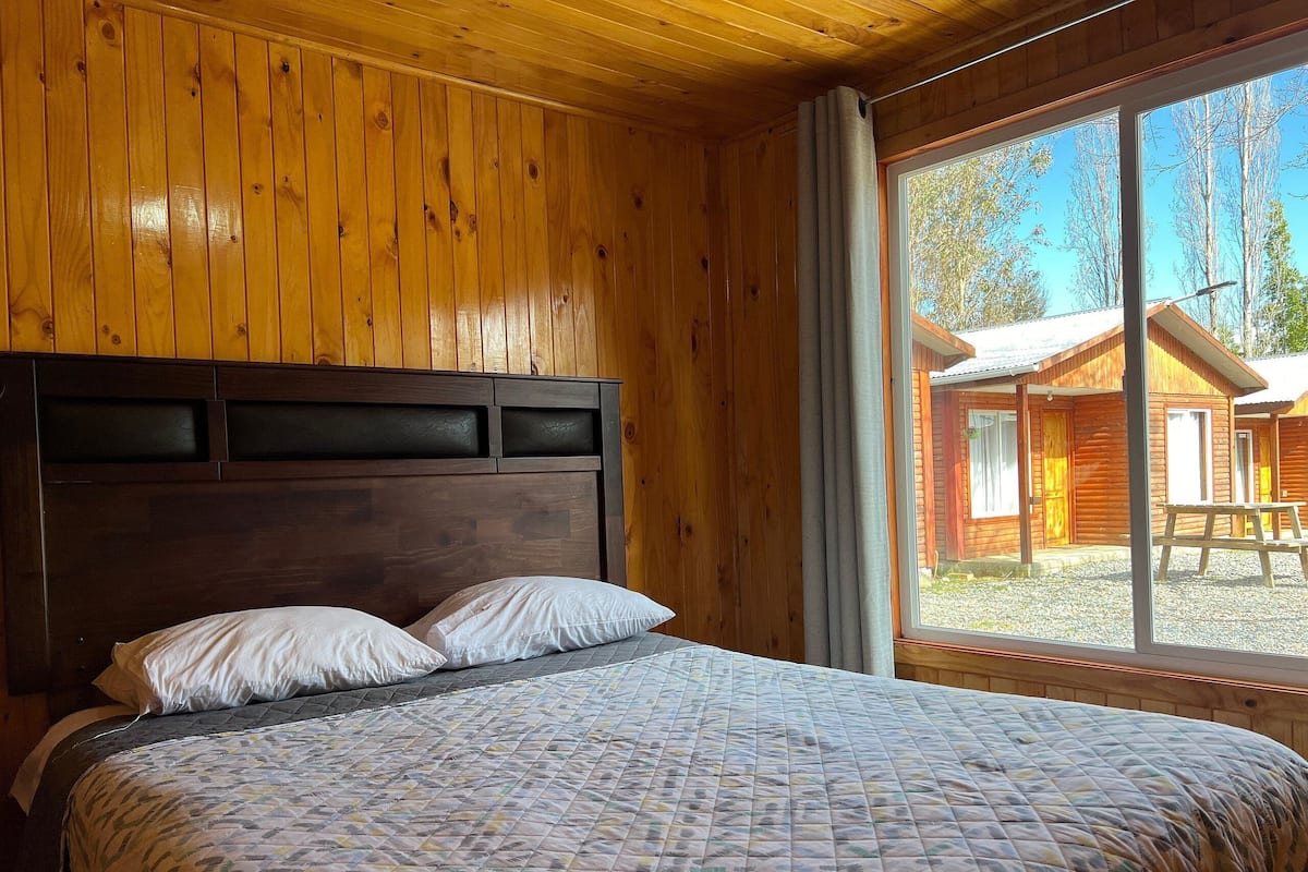 A wooden cabin room with a bed, a window, and a view of a cabin outside.