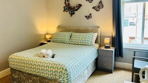 A neatly made bed with patterned bedding, two bedside lamps, a small table with a cup and snacks, and decorative butterfly wall art. - Le Papillon Hotel Blackpool (Blackpool)