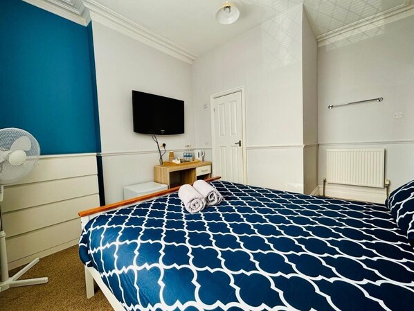 A bedroom with a bed, a TV, a bedside table, a fan, and a door. - Le Papillon Hotel Blackpool (Blackpool)