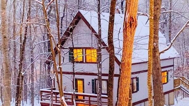 Uma cabana com deck de madeira e várias janelas, rodeada por árvores e neve.