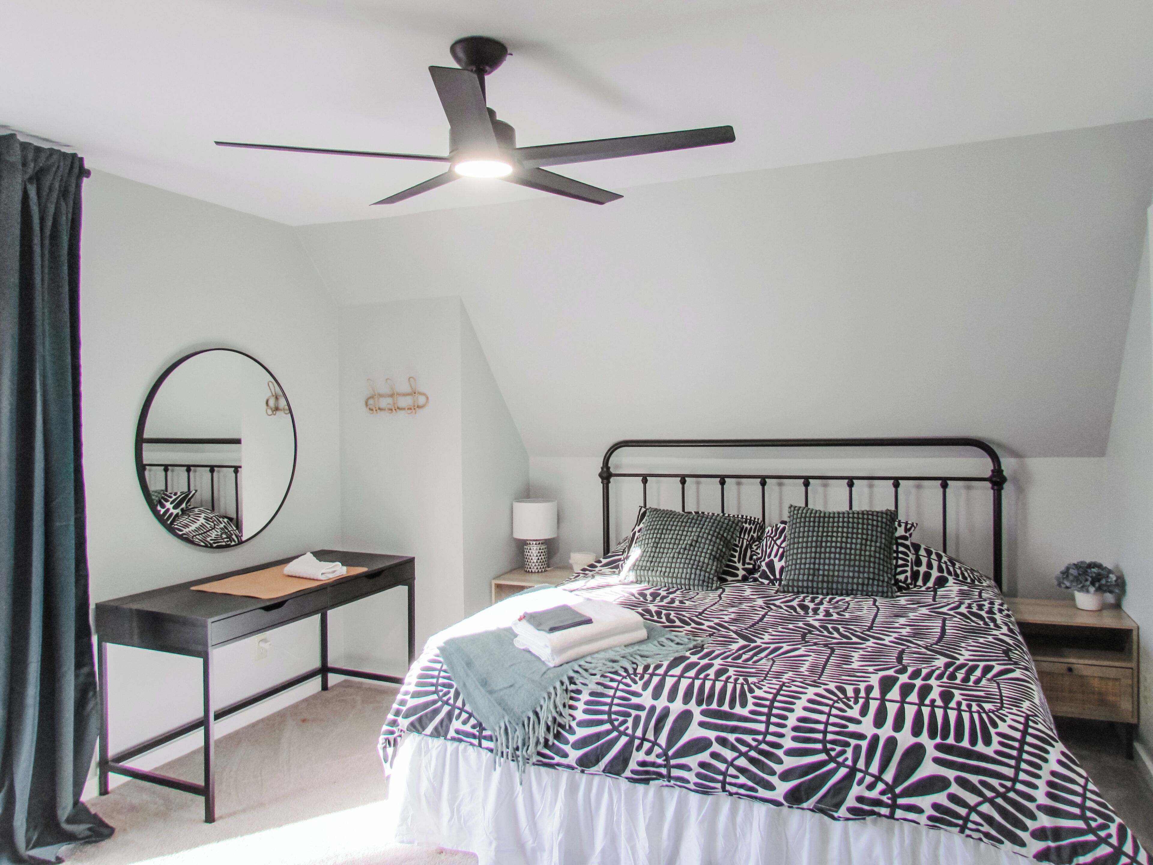 A bedroom with a bed, a desk, a chair, a ceiling fan, a mirror, and a nightstand.