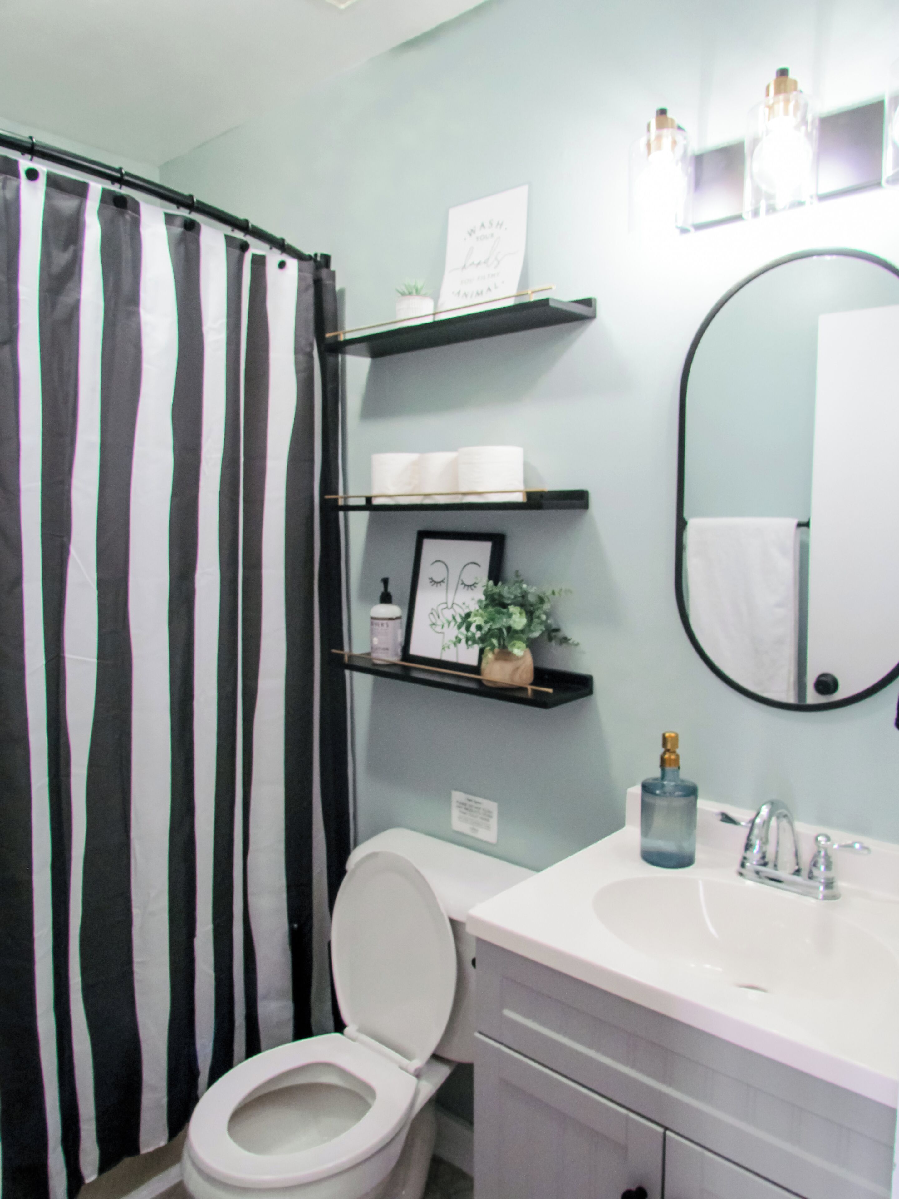 Un baño con cortina de ducha a rayas, un inodoro, un lavabo con espejo y estantes con artículos de aseo y un cuadro enmarcado.