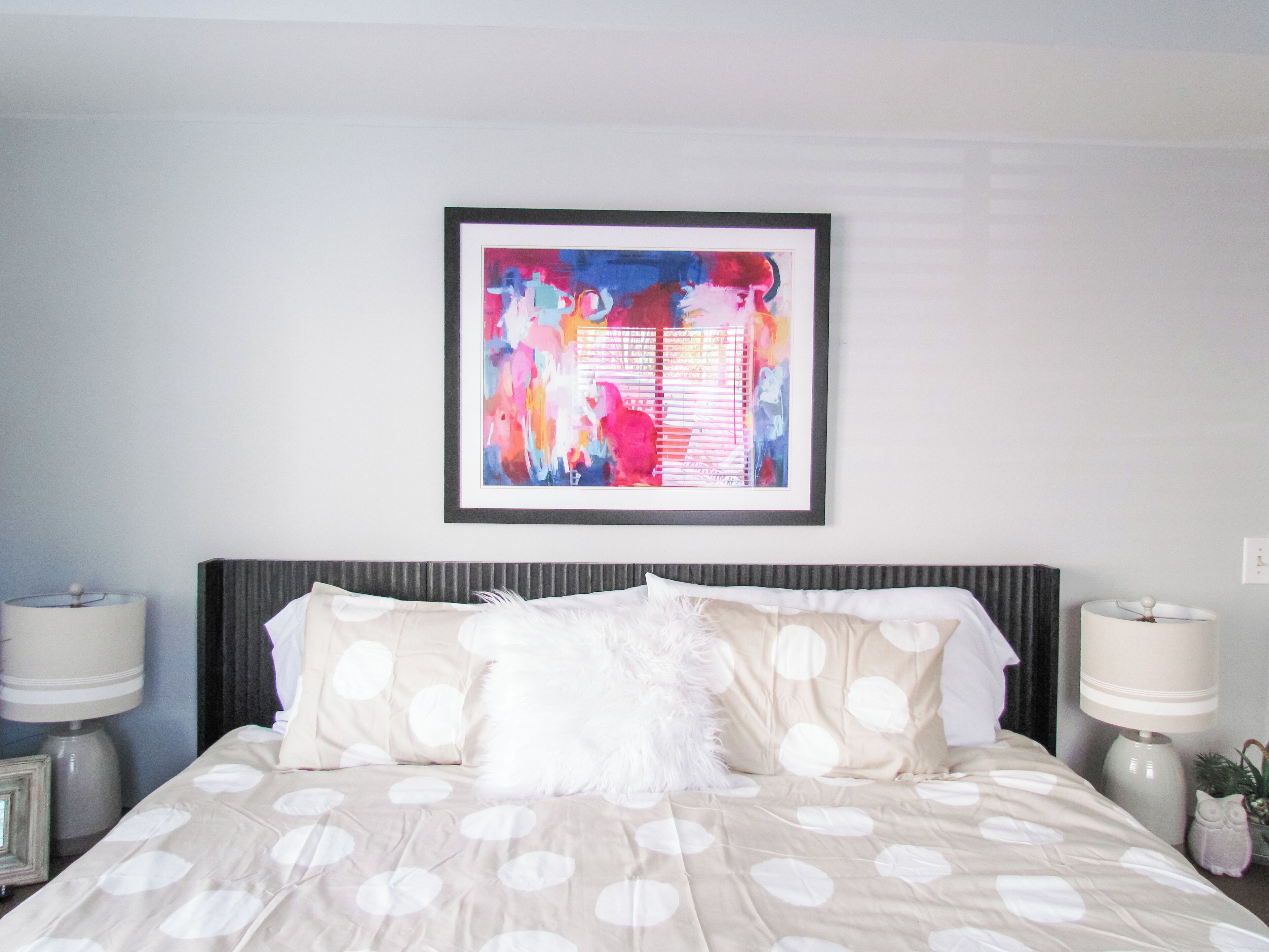 A neatly made bed with a polka dot bedspread, a framed abstract painting above it, and two bedside lamps on either side.