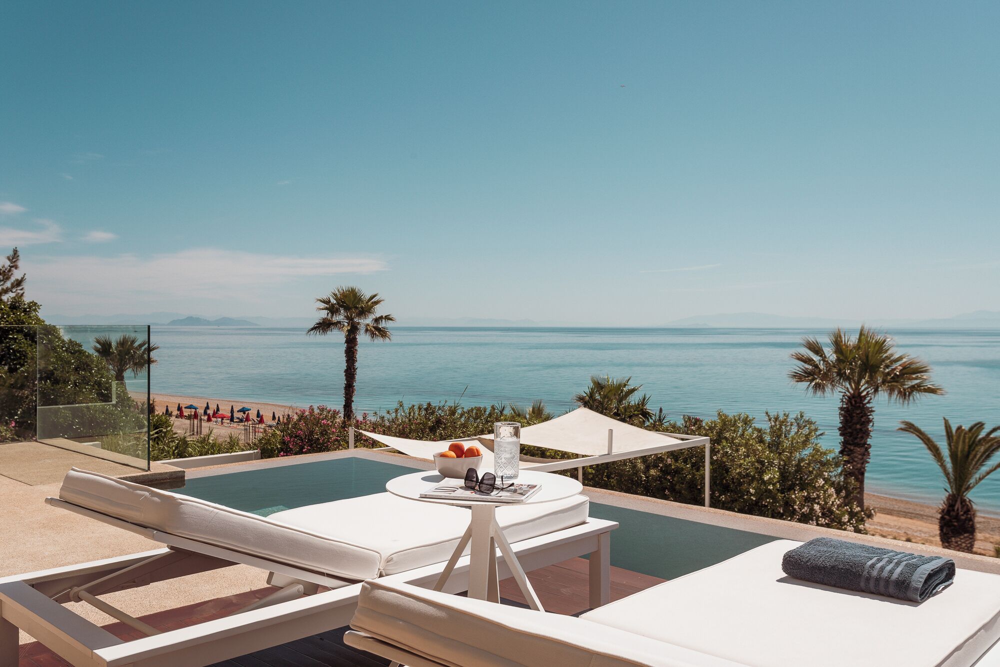 Un área junto a la piscina con sillones, una mesa con un frutero y vasos, y vista al mar.