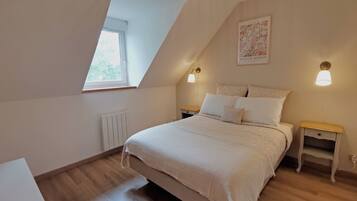 Un dormitorio con techo inclinado, una cama grande con ropa blanca, dos mesitas de noche con lámparas, un radiador y una ventana con vistas a árboles.