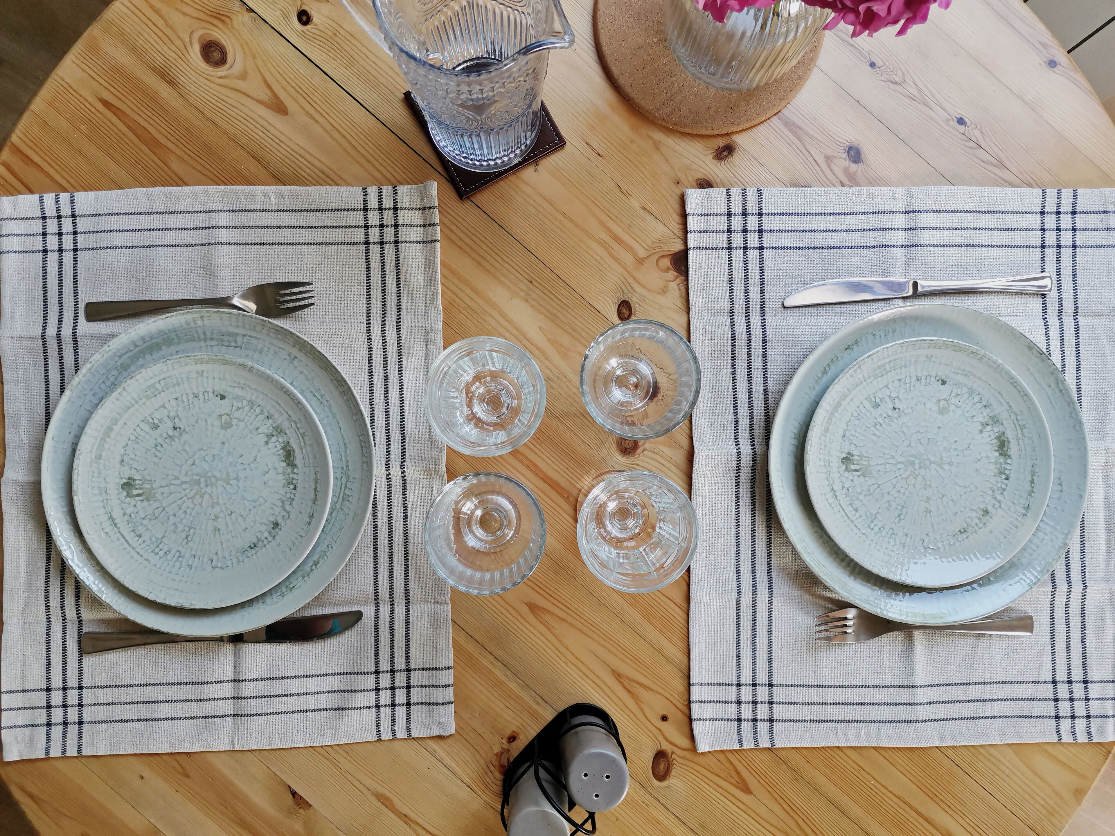 A wooden table set for two with two plates, two forks, two knives, and four clear glasses.