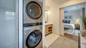 A modern bathroom with a double washing machine, a wooden vanity, a bed with a white and black patterned bedspread, and a small plant on the side. - Spacious 3bd Home~10min IU Baseball~2 K Beds~Huge Kitchen~Fire Pit+Ping Pong (Bloomington)