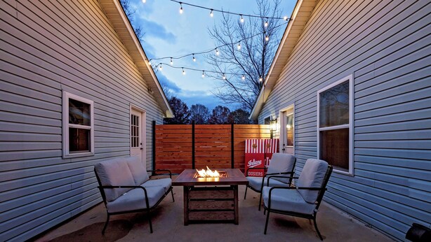 A patio with two chairs and a table with a fire feature, surrounded by wooden fencing and string lights. - Spacious 3bd Home~10min IU Baseball~2 K Beds~Huge Kitchen~Fire Pit+Ping Pong (Bloomington)