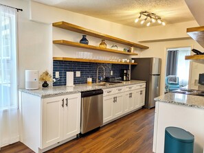 Una cocina moderna con armarios blancos, lavavajillas de acero inoxidable y horno empotrado. Hay repisas de madera sobre la encimera con diversos utensilios de cocina.