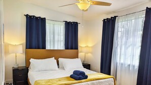 Un lit bien fait, avec du linge de lit blanc, une couverture jaune et une serviette bleue. Il y a deux tables de chevet avec des lampes, un ventilateur de plafond et une fenêtre avec des rideaux.