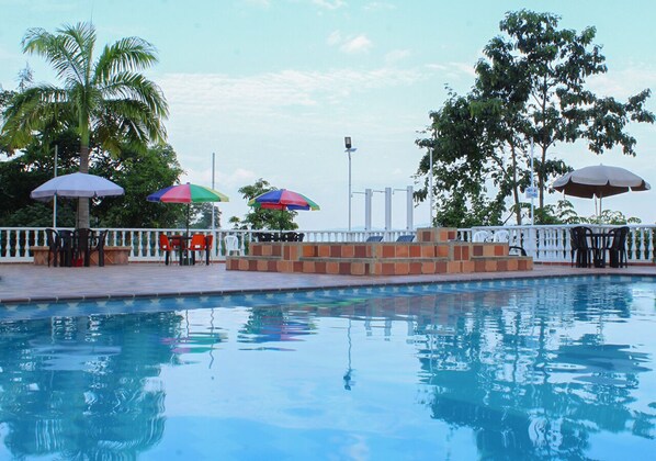 Piscina con sillas de descanso y sombrillas, rodeada de palmeras y una valla blanca.