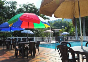 Zona de piscina al aire libre con varias mesas y sillas, sombrillas y una valla alrededor de la piscina.